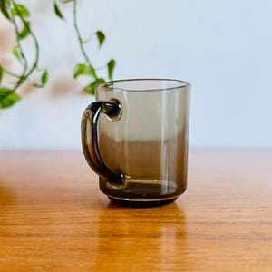 May include: A clear, brown-tinted glass mug with a rounded handle. The mug is cylindrical with a slightly flared base. The handle is a darker shade of brown. The mug is sitting on a wooden surface.