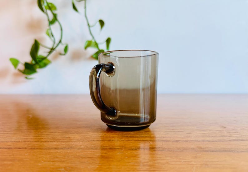 May include: A clear, brown-tinted glass mug with a rounded handle. The mug is cylindrical with a slightly flared base. The handle is a darker shade of brown. The mug is sitting on a wooden surface.