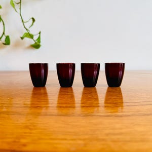 May include: Four small, dark red glasses are arranged in a row on a wooden surface. The glasses have a simple, rounded shape and a gradient colour, transitioning from a deep red at the top to a darker shade at the base. Green leaves are visible in the background.