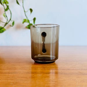 May include: A clear, brown-tinted glass with a unique handle design. The handle is a solid, dark brown shape that extends from the top to the bottom of the glass. The glass sits on a wooden surface.