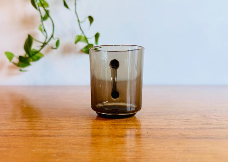 May include: A clear, brown-tinted glass with a unique handle design. The handle is a solid, dark brown shape that extends from the top to the bottom of the glass. The glass sits on a wooden surface.