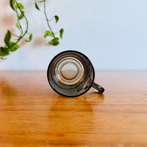 May include: A small, clear glass mug with a dark rim and handle. The mug is empty and sits on a wooden surface. Green leafy vines are visible in the background.