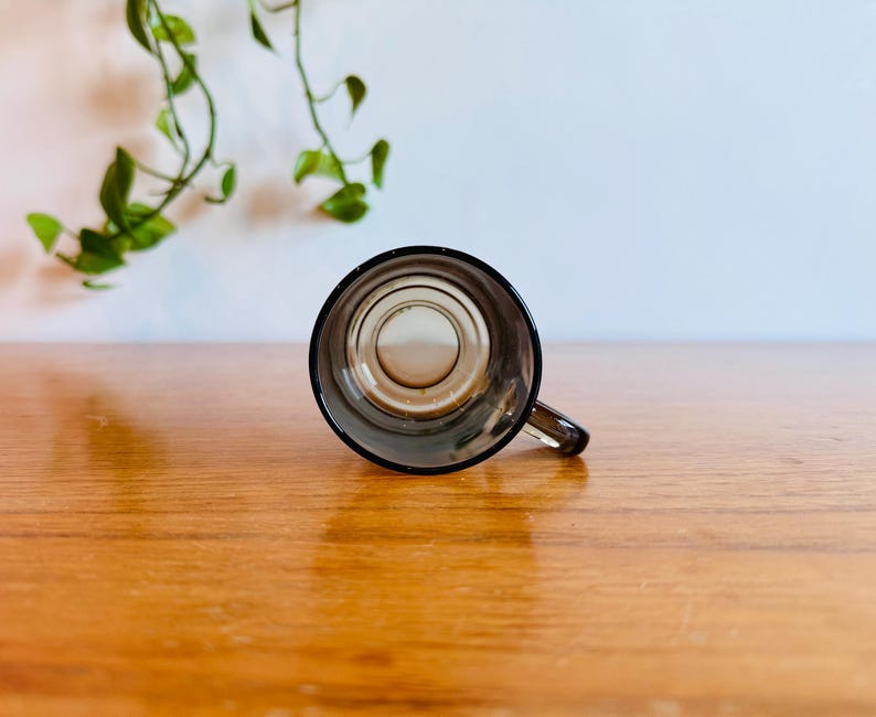 May include: A small, clear glass mug with a dark rim and handle. The mug is empty and sits on a wooden surface. Green leafy vines are visible in the background.