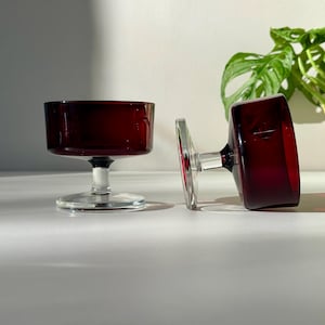May include: Two vintage ruby red glass coupe glasses. One glass is upright, the other is on its side. The glasses have a short stem and a wide, shallow bowl. The background is a white surface with a green plant.