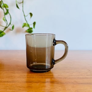 May include: A clear, brown-tinted glass mug with a rounded handle. The mug is cylindrical with a slightly flared base. The background includes a wooden surface and a white wall with green foliage.