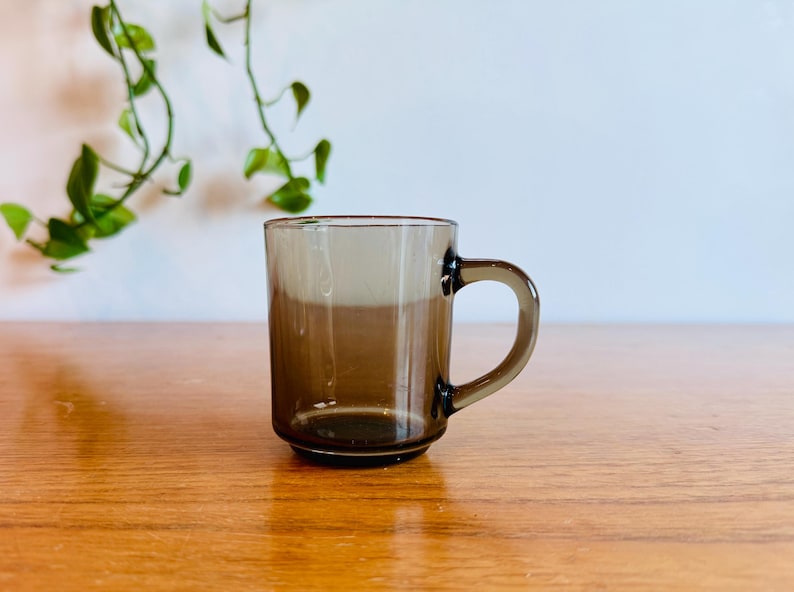 May include: A clear, brown-tinted glass mug with a rounded handle. The mug is cylindrical with a slightly flared base. The background includes a wooden surface and a white wall with green foliage.