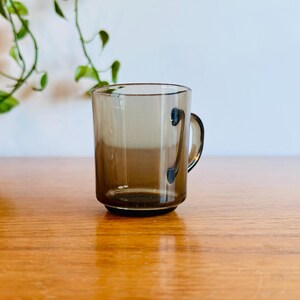 May include: A clear, brown-tinted glass mug with a handle. The mug is cylindrical with a slightly flared rim and sits on a wooden surface. A green plant is visible in the background.