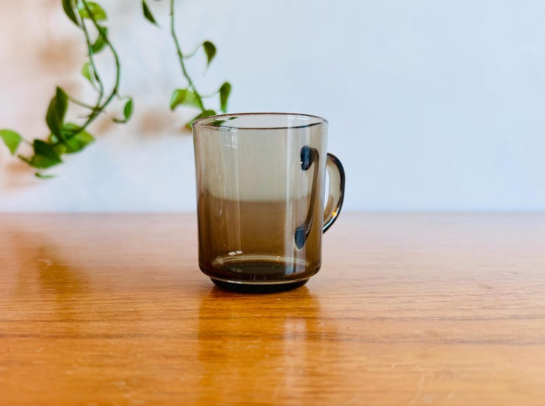 May include: A clear, brown-tinted glass mug with a handle. The mug is cylindrical with a slightly flared rim and sits on a wooden surface. A green plant is visible in the background.