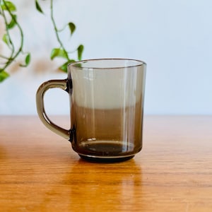 May include: A clear, brown-tinted glass mug with a handle. The mug sits on a wooden surface, with a plant in the background. The mug is approximately 3.5 inches tall.