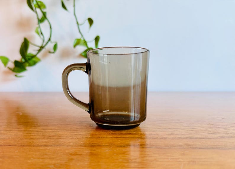 May include: A clear, brown-tinted glass mug with a handle. The mug sits on a wooden surface, with a plant in the background. The mug is approximately 3.5 inches tall.