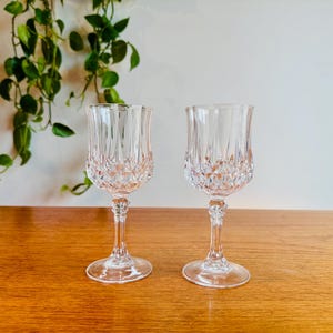 May include: Two clear crystal wine glasses with diamond-cut detailing. The glasses have a tulip shape and sit on a wooden surface. Green leafy vines hang in the background.
