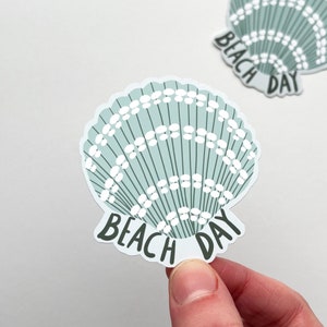 May include: Two seashell stickers in light teal and white. The stickers feature a scalloped edge and the words "BEACH DAY" in black text. The stickers are on a white background.