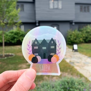 May include: A holographic sticker depicting a black house with three gables, pumpkins, and foliage within a snow globe design. The word "SALEM" is printed at the base. The sticker is held in front of a gray house.