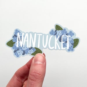 May include: A sticker with the word "NANTUCKET" in white, surrounded by blue hydrangea flowers and green leaves. The sticker has a light blue background and is held by a hand.