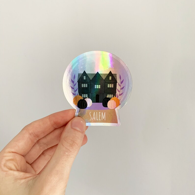 May include: Holographic sticker with a snow globe design. It features a dark house, pumpkins, and the word "SALEM". The sticker has an iridescent finish. The design includes a house with multiple windows and pumpkins in various colours.