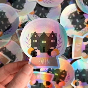 May include: Holographic sticker depicting a black house with three gables, pumpkins, and foliage within a snow globe design. The word "SALEM" is printed at the base. The sticker is held by a hand.