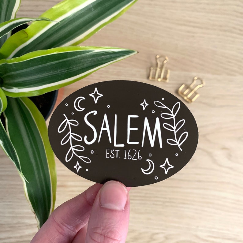 May include: Black oval sticker with the word "SALEM" in white, surrounded by stars, moons, and leaves. The text "EST. 1626" is below the main text. The sticker is held in a hand near a plant and gold binder clips.