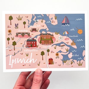 May include: A colorful illustrated map of Ipswich, Massachusetts, featuring landmarks like the Hall-Haskell House and Crane Beach. The map is in a whimsical style with a pink and blue color scheme, and the town's name is written in a cursive font.
