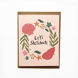 May include: Greeting card with a pink background and a brown border. The card features a wreath of seashells, starfish, and flowers. The text "Let's Shellebrate" is written in black script.