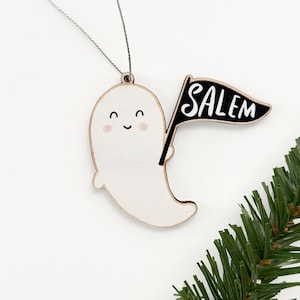 Peut inclure: Ornement en bois en forme de fant&ocirc;me souriant, tenant un drapeau noir avec l'inscription "SALEM" en blanc. L'ornement est blanc avec un bord en bois naturel et une ficelle argent&eacute;e pour l'accrocher. Une branche de pin vert est en arri&egrave;re-plan.