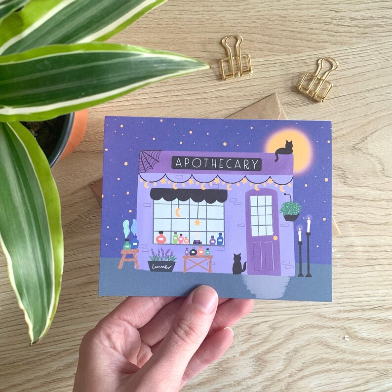 May include: Apothecary illustration on a greeting card. The purple building has a cobweb, string lights, and a sign that reads "APOTHECARY". A black cat sits on the roof under a full moon. The card is held by a hand.