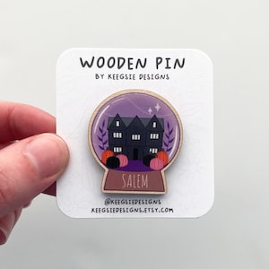 May include: Wooden pin featuring a black house inside a purple snow globe with the word "Salem" below. The pin includes pumpkins and the text "WOODEN PIN BY KEESGIE DESIGNS" and the Etsy shop URL.
