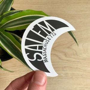 May include: A black and white sticker in a crescent moon shape. The word "SALEM" is printed in large, bold, white letters, with "MASSACHUSETTS" printed below in smaller white letters. The sticker is held in front of a green and white plant.