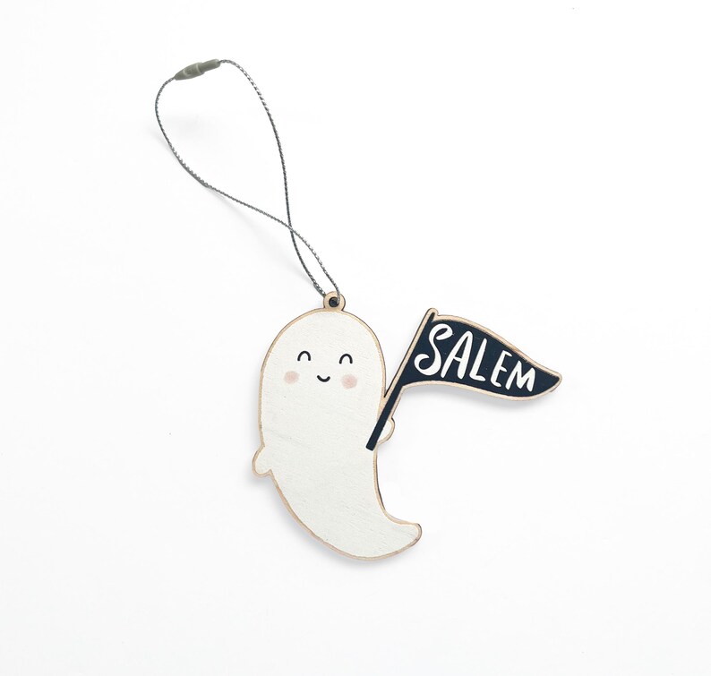 Peut inclure: Ornement en bois en forme de fant&ocirc;me souriant, tenant un drapeau noir avec l'inscription "SALEM". L'ornement est dot&eacute; d'une ficelle pour &ecirc;tre suspendu. Le fant&ocirc;me est blanc avec un grain de bois clair.