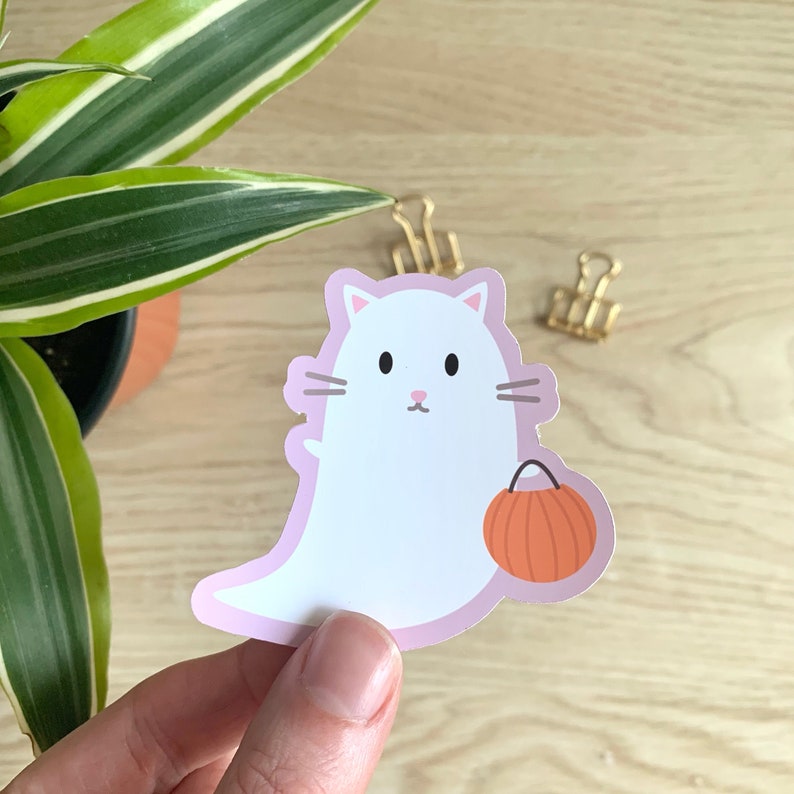 May include: A white cat ghost sticker with a pink outline, holding an orange pumpkin-shaped candy bag. The sticker has black eyes, whiskers, and a pink nose. A Halloween-themed decorative item.