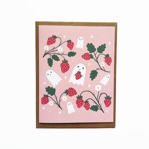 Puede incluir: Una tarjeta de felicitación con un fondo rosa que presenta un diseño caprichoso de fresas, hojas verdes, flores blancas y personajes de fantasmas amigables. La tarjeta está enmarcada en un marco de cartón marrón.