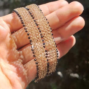 May include: Gold-coloured chain bracelets with black beads. The bracelets are made of delicate, interwoven chains, creating a textured surface. The black beads are evenly spaced along the edges, adding a contrasting detail. The bracelets are displayed against a blurred green background.