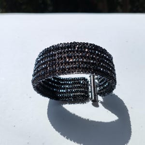 May include: A wide, black beaded bracelet with a silver-tone clasp. The bracelet is made of small, dark beads woven together in a wide band. The clasp is a simple, straight bar that inserts into a loop. The bracelet casts a shadow on a white surface.
