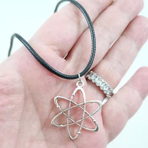 May include: A silver atom pendant necklace on a black cord. The pendant is a stylized representation of an atom with a central nucleus and orbiting electrons.