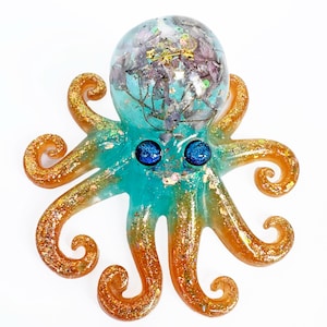Puede incluir: Figura artesanal de un pulpo con cuerpo turquesa y tentáculos naranja dorado. La cabeza es una cúpula transparente llena de flores secas y purpurina. El pulpo tiene dos ojos azules brillantes, sobre un fondo blanco.
