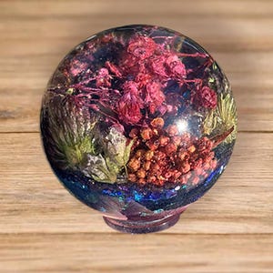 May include: A clear spherical resin paperweight containing preserved dried flowers in shades of deep pink, red, and green, with small orange berries and iridescent blue glitter flakes embedded within the transparent material at the base.
