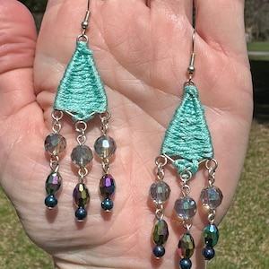 May include: A pair of turquoise blue dangle earrings with iridescent glass beads. The earrings are made with woven thread and have a triangular shape.
