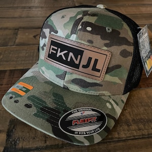 Può includere: Un cappello da camionista mimetico con rete nera sul retro. Il cappello ha una toppa in pelle marrone con il testo "FK NJL" in lettere bianche.