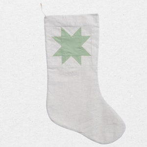 May include: A light gray fabric Christmas stocking featuring a sage green eight-pointed star appliqué centered on the upper section. The stocking has a simple, clean design with a small fabric loop attached to the top corner for hanging purposes.
