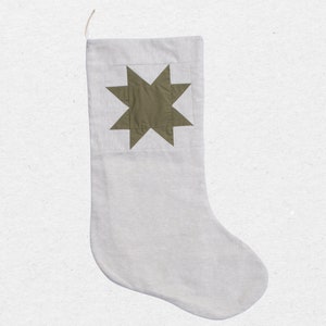 May include: A light gray holiday stocking featuring a centered olive green eight-pointed star patch on the upper section. The stocking is constructed from a textured fabric and includes a small fabric loop at the top corner for hanging purposes.