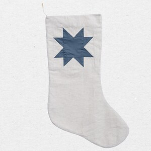 May include: A light gray fabric Christmas stocking featuring a centered, dark blue eight-pointed star appliqué. The stocking has a simple, flat construction with a small fabric loop attached to the top left corner for hanging purposes.