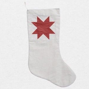 May include: A light gray fabric Christmas stocking featuring a large, eight-pointed star applique in a deep red color centered on the upper section. The stocking includes a small, thin fabric loop attached to the top corner for hanging purposes.