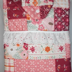 Puede incluir: Una funda de tela acolchada con un diseño de patchwork de patrones florales y de cuadros en tonos rosa, rojo y blanco. La funda incluye una solapa con un cierre de presión blanco y un borde de encaje blanco con ojales en el centro.