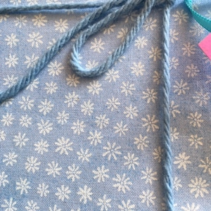 May include: Close-up of a light blue yarn tied in a bow around a package. The package is wrapped in light blue fabric with a white floral pattern. A small pink tag is attached to the bow.