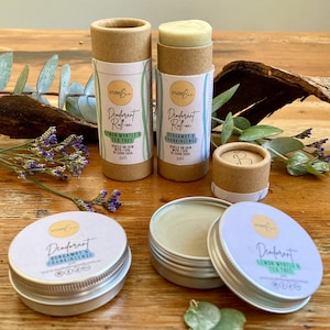 May include: Four natural deodorant containers, two in cardboard tubes and two in metal tins. The tubes are labelled "Deodorant Roll-on" with the scents "Lemon Myrtle & Tea Tree" and "Bergamot & Frankincense". The tins are labelled "Deodorant" with the same scents. All containers are on a wooden surface with green foliage.