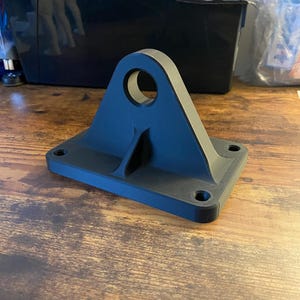 May include: A matte black 3D printed mounting bracket featuring a rectangular base with four circular screw holes at the corners and a central vertical support fin with a large circular opening at the top for mechanical attachment purposes.