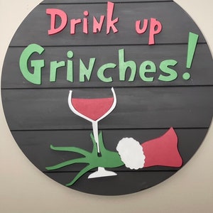 May include: A round wooden sign with the text "Drink up Grinches!" in green and red letters. A green hand holding a glass of red wine is depicted below the text.