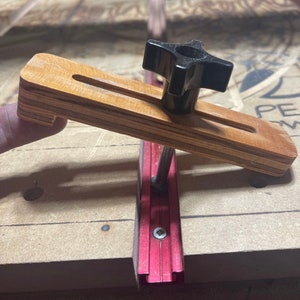May include: A wooden clamp with a black knob, attached to a red aluminum track. The clamp is used to secure a piece of wood to the track.