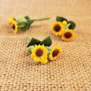 May include: Three small artificial sunflowers with green leaves on a burlap background. The sunflowers are yellow with brown centers.