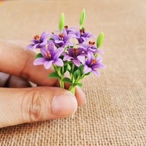 以下が含まれることがあります： 緑色の茎を持つ、紫と白の人工の花のブーケ。花は手に持たれています。