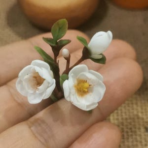 May include: A small bouquet of white flowers with yellow centers, crafted from polymer clay. The flowers are arranged on a branch with green leaves.